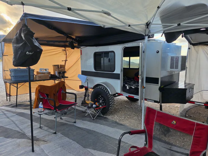 An RV rental camping site with a silver teardrop trailer, canopy, chairs, and camping gear on a striped mat, set against mountains.. Runaway Cool Camp 4x8 2018