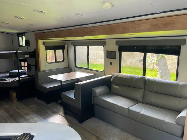 RV rental interior showing a grey sofa, booth dinette, and bunk beds. Bright windows illuminate the living and dining areas.. Keystone RV Springdale 2022