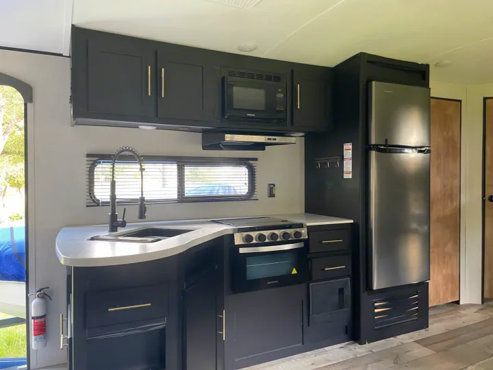 This modern rv rental kitchen boasts dark cabinets, a stainless fridge, microwave, stove, and sink. Compact & functional.. Keystone RV Springdale 2022