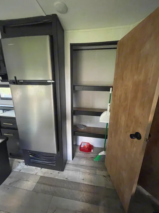 Inside an RV rental: a compact stainless steel fridge, shelved pantry with cleaning supplies, and wood-look floor.. Keystone RV Springdale 2022