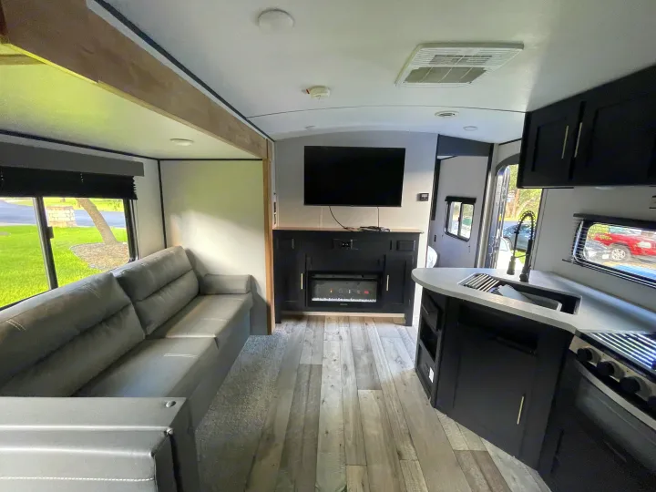 Modern rv rental interior featuring a gray couch, TV, fireplace, and kitchen area. Perfect for comfortable travel.. Keystone RV Springdale 2022