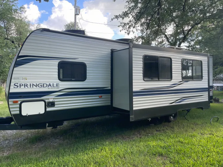 This Springdale Keystone travel trailer rv rental is parked outdoors with its slide-out extended, surrounded by grass and trees.. Keystone RV Springdale 2022