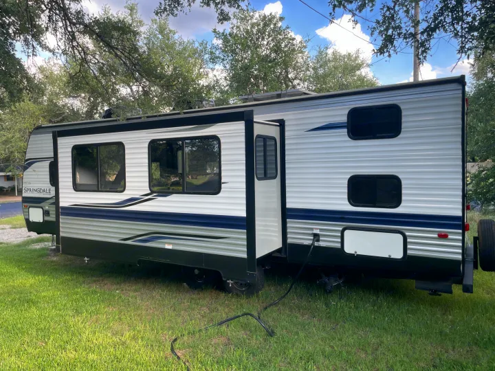 This Springdale travel trailer, likely for rv rental, is parked on grass with its slide-out extended, showing power & sewage hookups.. Keystone RV Springdale 2022