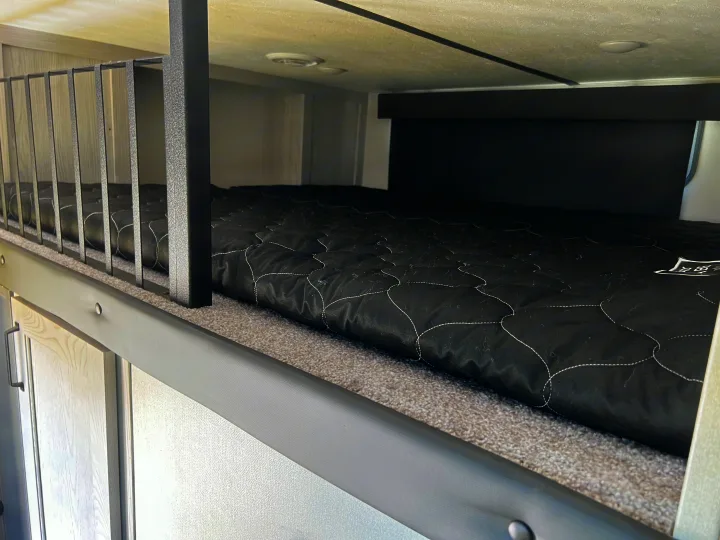 Close-up of a cozy RV rental bunk bed interior, featuring a black quilted mattress, metal safety rail, and carpeted sleeping area.. Keystone RV Montana 2022