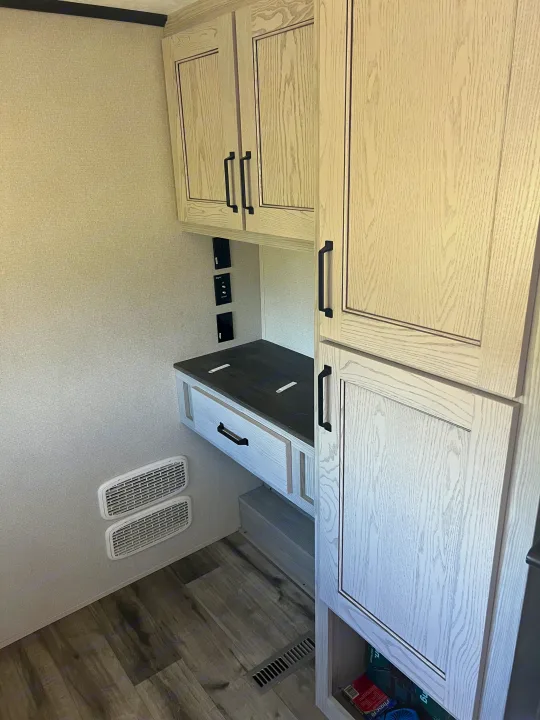 RV rental interior: light wood-grain cabinets, a compact desk area with power outlets, textured walls, and wood-look flooring.. Keystone RV Montana 2022