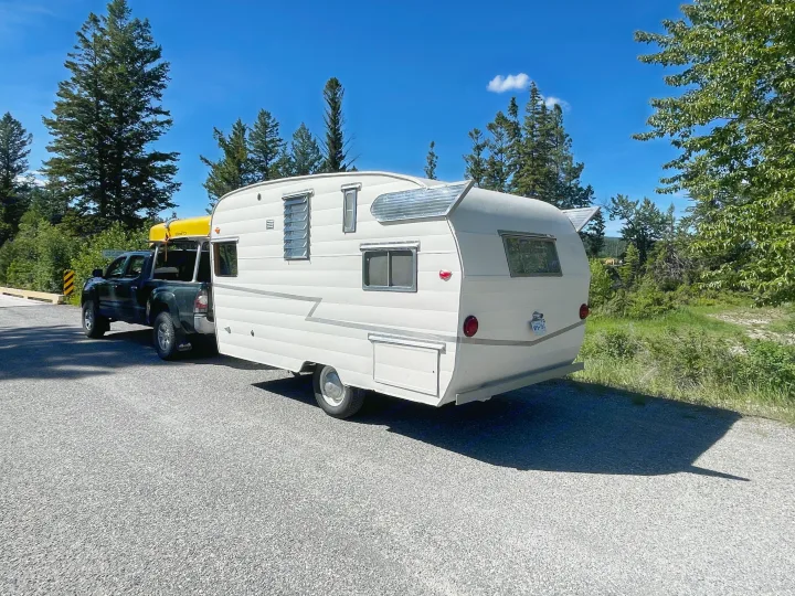 Explore with an RV rental! Vintage travel trailer & truck with a canoe ready for a sunny road trip amidst tall green trees.. Shasta Airflyte 1958