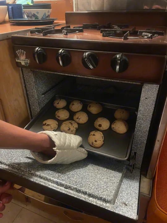 Hand in oven mitt places chocolate chip cookies on a tray into a Princess RV rental oven. Fresh baked goods on the road!. Shasta Airflyte 1958