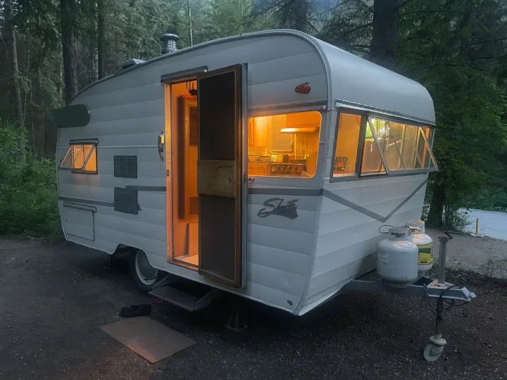Cozy vintage Shasta RV rental glows at dusk in the forest. Its open door and windows reveal a warm, inviting interior.. Shasta Airflyte 1958
