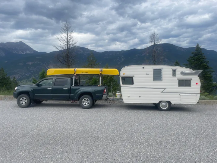 Truck with canoe towing a retro RV rental, parked against a scenic mountain backdrop under cloudy skies.. Shasta Airflyte 1958