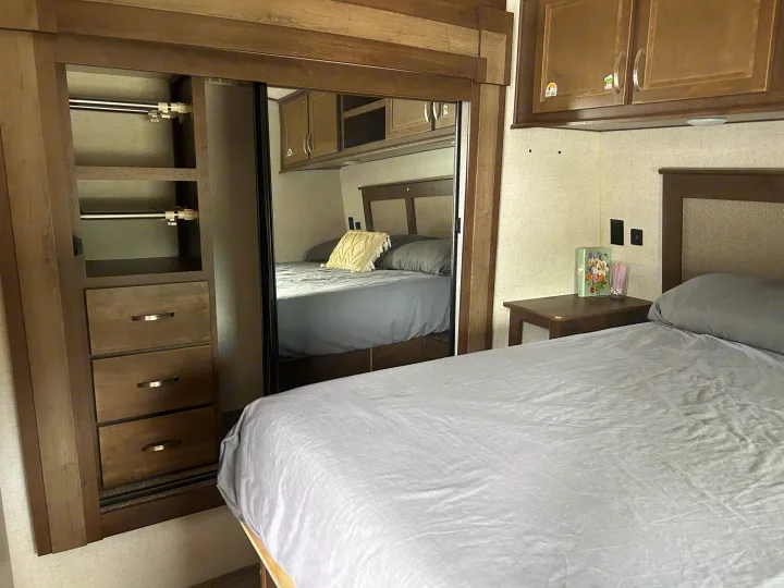 RV rental bedroom with a gray bed, bedside table, mirrored closet, and wood cabinets for storage.. Highland Ridge RV Open Range 2020