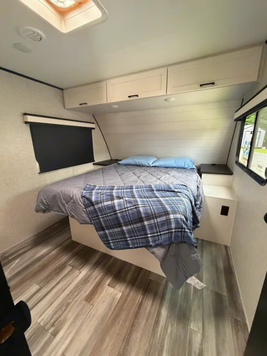 Cozy RV rental bedroom with a gray bed, blue plaid blanket, light blue pillows, overhead cabinets, and wood-look floor.. Jayco Eagle HT 2025
