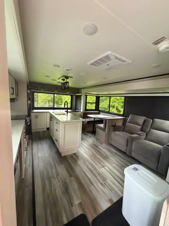 Modern RV rental interior showing kitchen island, dinette, and gray recliners on wood-look floor, with outdoor views.. Jayco Eagle HT 2025