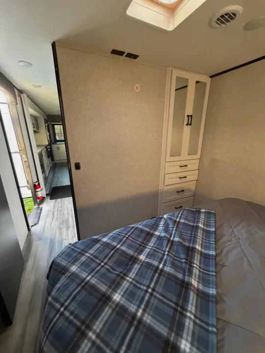 Cozy RV rental bedroom featuring a blue plaid blanket, built-in cabinet with drawers, and an open view to the mobile living space.. Jayco Eagle HT 2025