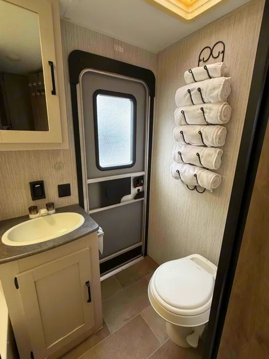 RV rental bathroom: compact sink, toilet, door with frosted window, and a wall-mounted rack of rolled towels. Exit sign visible.. Forest River Palomino Puma XLE Lite 2021