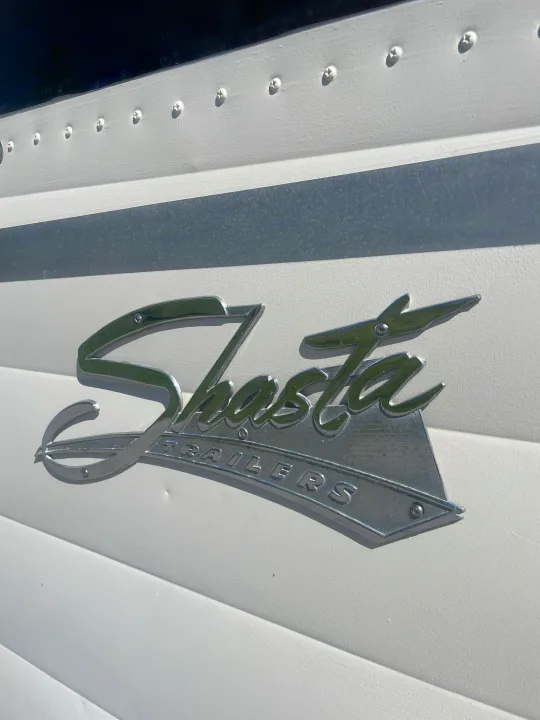 Close-up of a shiny Shasta Trailers emblem on a vintage RV rental. White textured siding with visible screws and metallic badge details.. Shasta Airflyte 1958