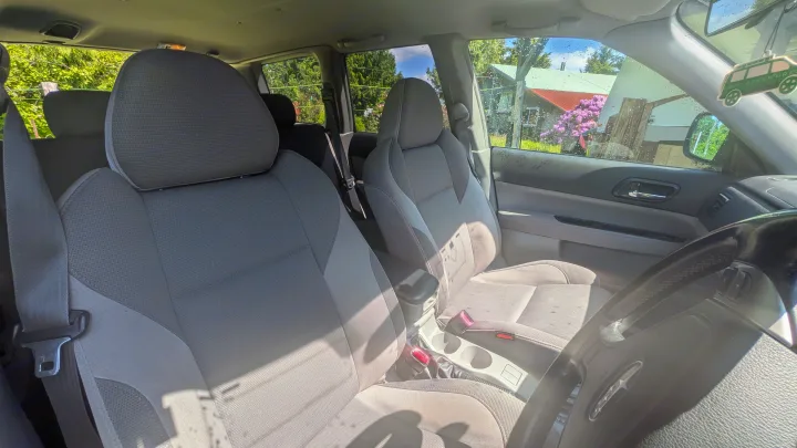Inside a car, gray front seats and steering wheel visible. An RV rental-style air freshener hangs, with trees and a house outside.. Subaru Forester 2009