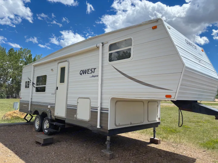 White QWEST by Jayco fifth-wheel RV rental, parked on gravel with trees and blue sky. Features an awning and storage. Ready for adventure.. Jayco Qwest 2003