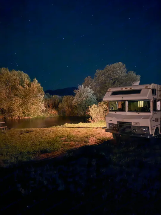 A white RV is parked at a scenic, starlit rv rental campsite by a pond, surrounded by illuminated trees at night.. International 510A 1973