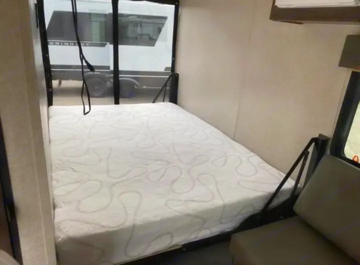 Compact RV rental bedroom with a comfy white patterned mattress. Window view shows another RV outside, next to a seating area.. Sunset Park RV SunRay 2022