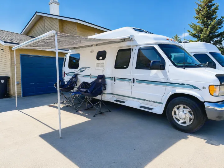 A white camper van, an RV rental, with an awning extended over two chairs on a driveway next to a house.. Pleasure-Way Pleasure-Way Class B 2002