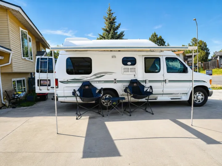 White camper van (rv rental) with extended awning, two camping chairs & table on a driveway next to a house.. Pleasure-Way Pleasure-Way Class B 2002