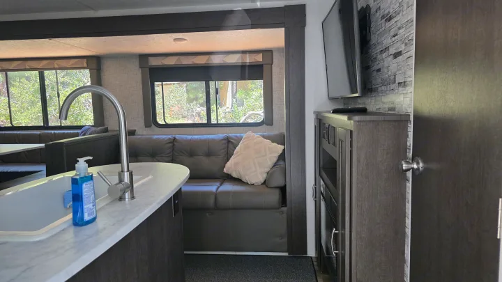 Spacious rv rental interior: modern kitchen with sink, dark living area sofa, TV, and windows with forest views.. Forest River Wildwood X-Lite 2019