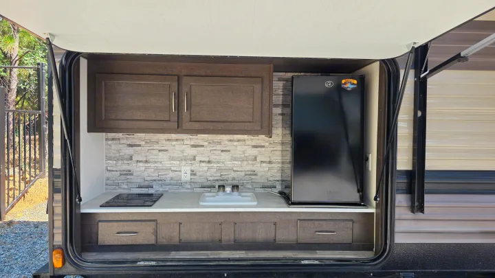 This RV rental's outdoor kitchen features a sink, cooktop, mini-fridge, and storage cabinets, ready for your camping adventures.. Forest River Wildwood X-Lite 2019