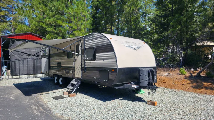 Wildwood travel trailer rv rental with an extended awning, parked on gravel near a red shed and tall pine trees.. Forest River Wildwood X-Lite 2019