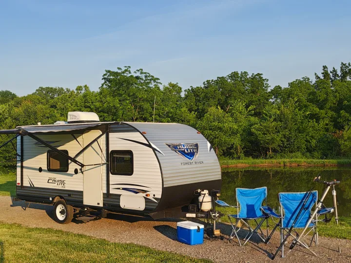An RV rental sits by a calm lake with blue camping chairs, fishing rods, and a cooler, ready for a serene outdoor escape.. Forest River Salem FSX 2018