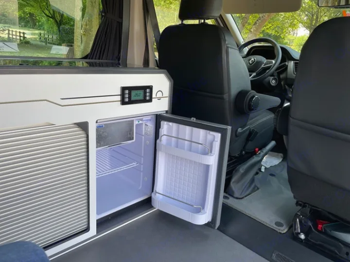 Inside an rv rental: an open mini-fridge displaying 26.0 on the control panel, part of a kitchenette unit, and the driver's cabin.. HANROAD TREK 5 2026