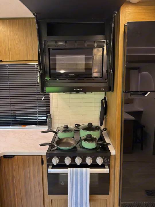 This RV rental kitchen features a black microwave, range hood, gas stove with green pots/pans, and refrigerator. Ideal for road trip cooking.. Forest River Wildwood FSX Platinum 2026