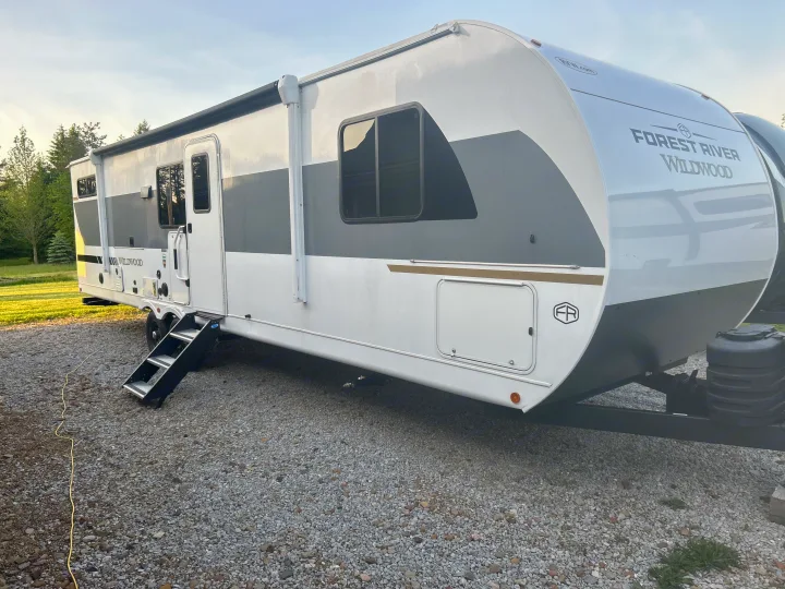 White and gray Forest River Wildwood rv rental travel trailer parked on gravel with steps down. Trees in the background.. Forest River Wildwood FSX Platinum 2026