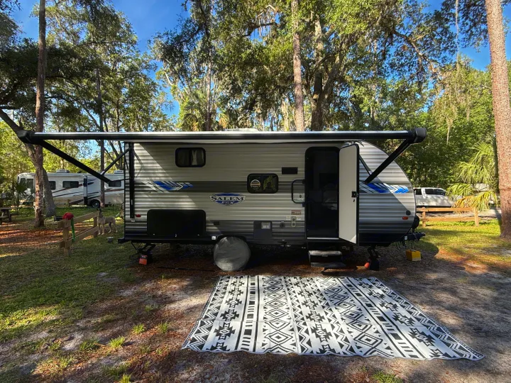 RV rental travel trailer parked at a shaded campsite with an extended awning, outdoor mat, and a dog enjoying nature.. Forest River Salem FSX 2022