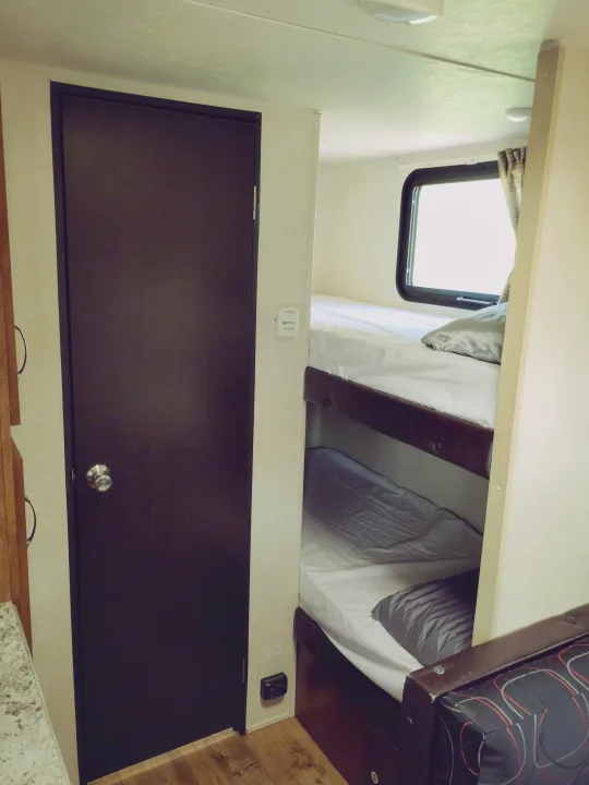 RV rental interior features a dark door, thermostat, and cozy bunk beds with clean white sheets and gray pillows. Includes safety detector.. Forest River Salem FSX 2017