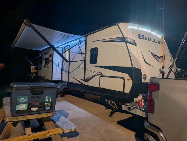 This illuminated rv rental (Keystone Bullet) is set up for a night of camping. A cooler and fishing gear are ready for outdoor adventures.. Keystone RV Bullet Ultra Lite 2023