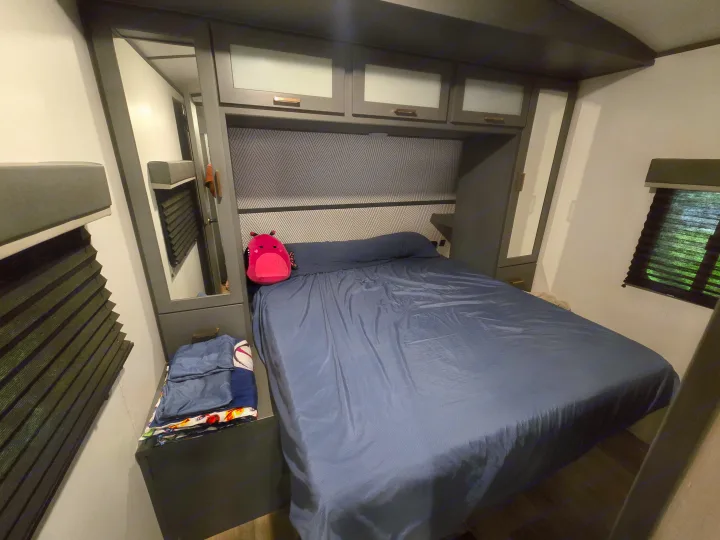 Cozy rv rental bedroom with a comfy bed, blue sheets, overhead storage, and mirrored wardrobes. A pink plushie adds charm.. Keystone RV Bullet Ultra Lite 2023