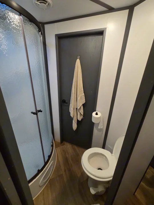 Compact RV rental bathroom with curved glass shower, toilet, and a towel hanging on the dark door.. Keystone RV Bullet Ultra Lite 2023