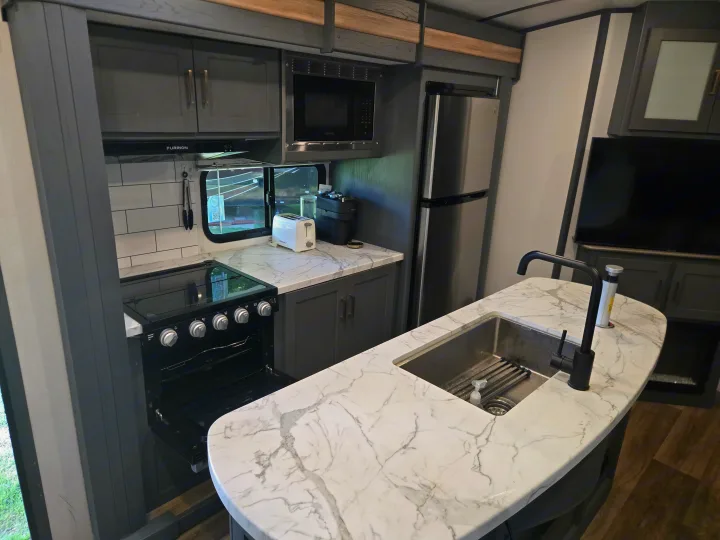 Spacious RV rental kitchen with modern dark cabinets, marble counters, stove, microwave, fridge, and an island sink.. Keystone RV Bullet Ultra Lite 2023