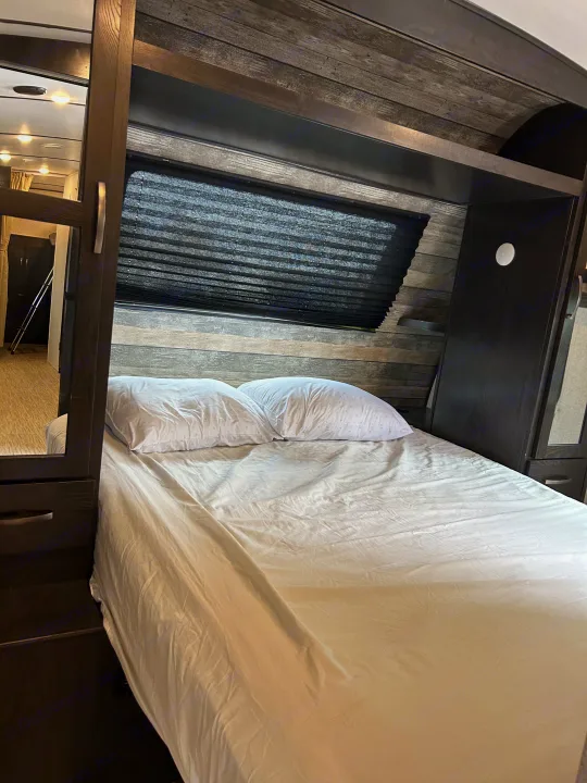 Relax in this RV rental bedroom, boasting a comfy bed with white linens, a textured wood-paneled wall, and a window with blinds.. Surveyor Legend 2021