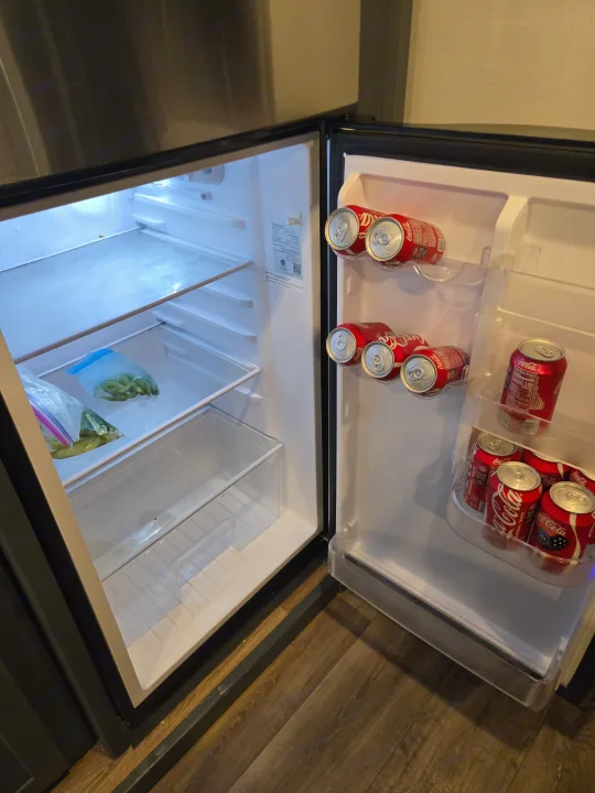An open mini-fridge in an RV rental, stocked with Coca-Cola cans and bags of green vegetables.. Keystone RV Bullet Ultra Lite 2023