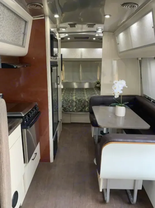 This RV rental interior features a kitchen with stove, a dining booth with a plant, and a rear bedroom with patterned bedding.. Airstream International 2017