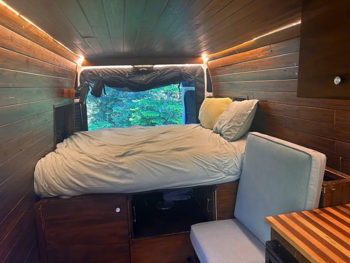 Cozy RV rental interior with wood-paneled walls, bed, ambient strip lighting, window view of trees, cushioned seat, & storage.. RAM Promaster 2022
