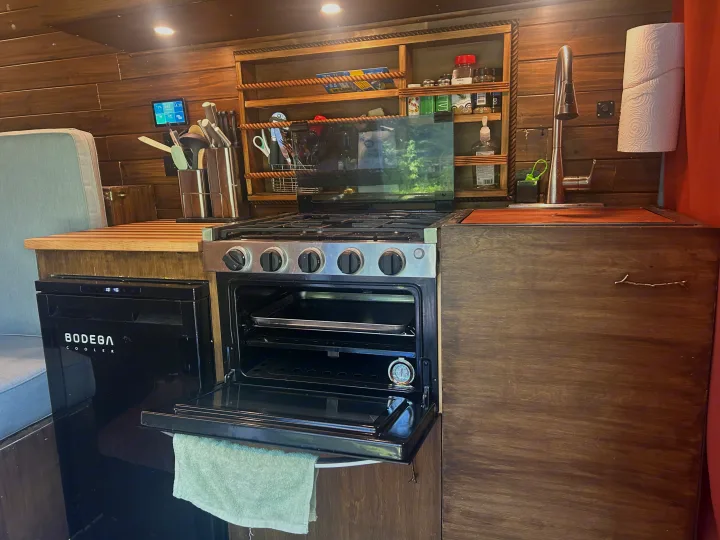 Inside an RV rental, a compact kitchen features a gas stove/oven, sink, and Bodega cooler amidst rustic wooden finishes.. RAM Promaster 2022