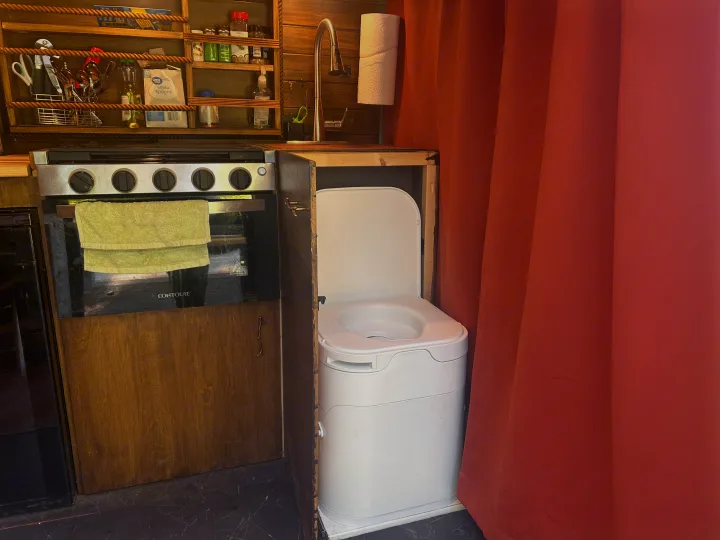 RV rental: Compact kitchen with stove, sink, and shelves next to a portable toilet, partially hidden by a red privacy curtain.. RAM Promaster 2022