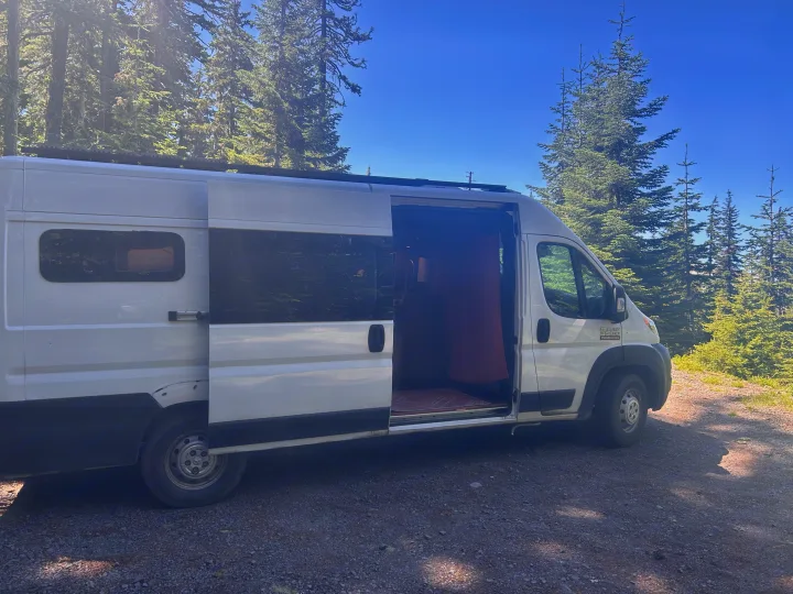 White RV rental camper van with an open door, parked on a sunny gravel path among tall evergreen trees under a clear blue sky.. RAM Promaster 2022