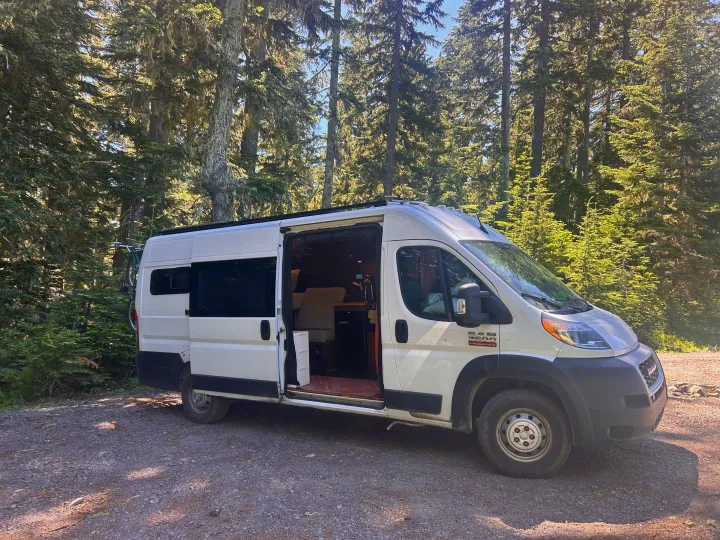White camper van (RV rental) parked in a sunny forest with its side door open, revealing the interior. A bike is mounted on the back.. RAM Promaster 2022