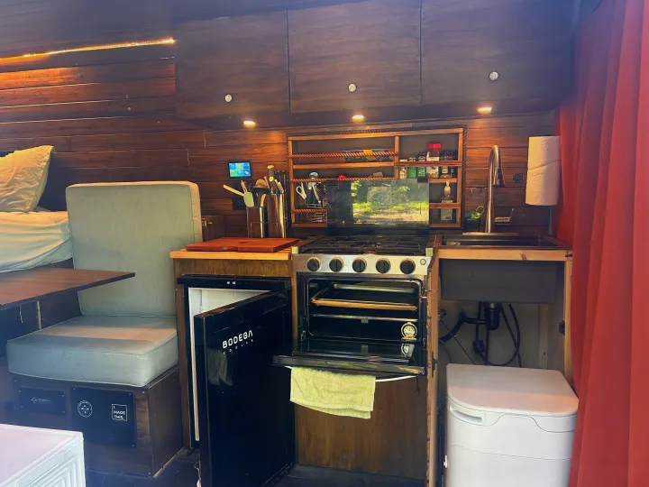 This RV rental's cozy interior features a full kitchenette with stove, oven, fridge, sink, a dinette, and a bed, all in warm wood tones.. RAM Promaster 2022