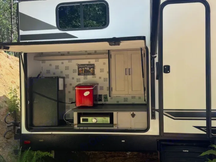 This rv rental boasts an exterior kitchen with a mini-fridge, red cooler, grill, and cabinets, ideal for outdoor camping.. Heartland RVs Mallard 2023