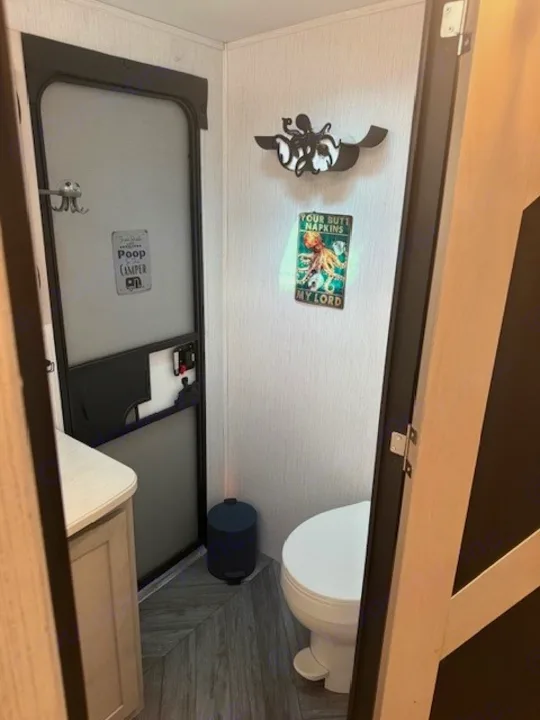 RV rental bathroom with toilet, trash can, vanity, and humorous signs like "Your Butt Napkins My Lord.". Heartland RVs Mallard 2023