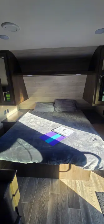 Interior of an RV rental bedroom with a grey bed, overhead cabinets, and sunlit wood-look floor.. Jayco Jay Flight SLX 2021
